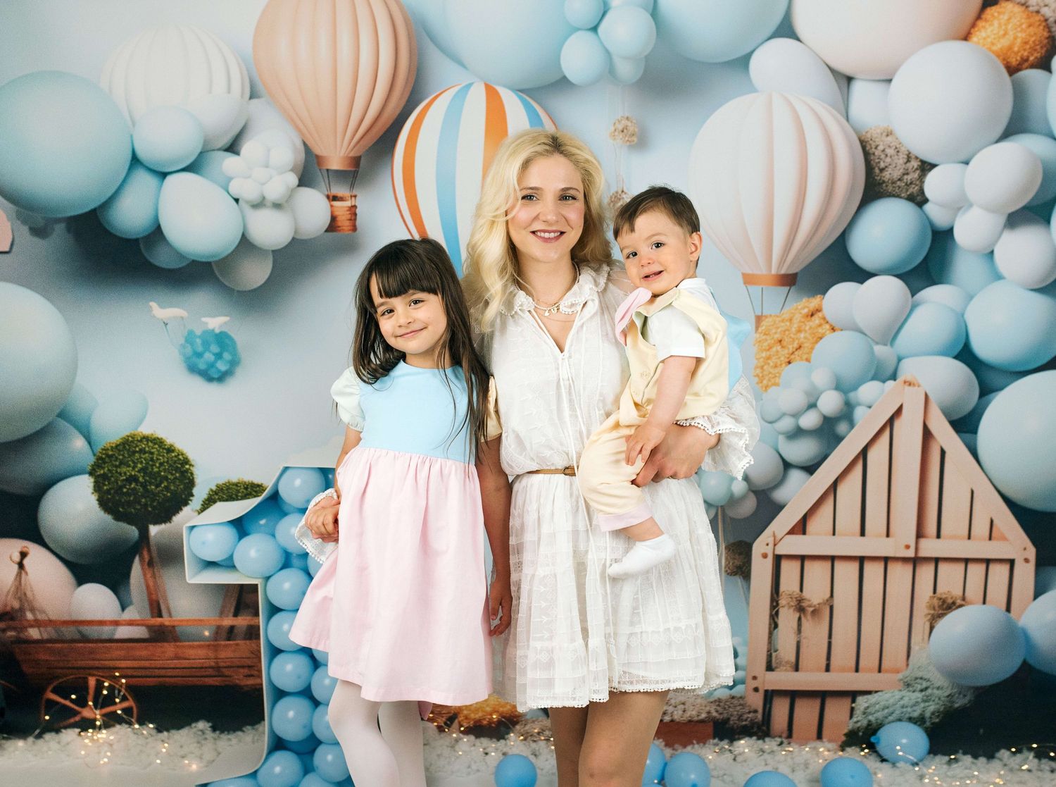 Woman in white dress holds baby while posing with young girl in pink dress before balloon backdrop.