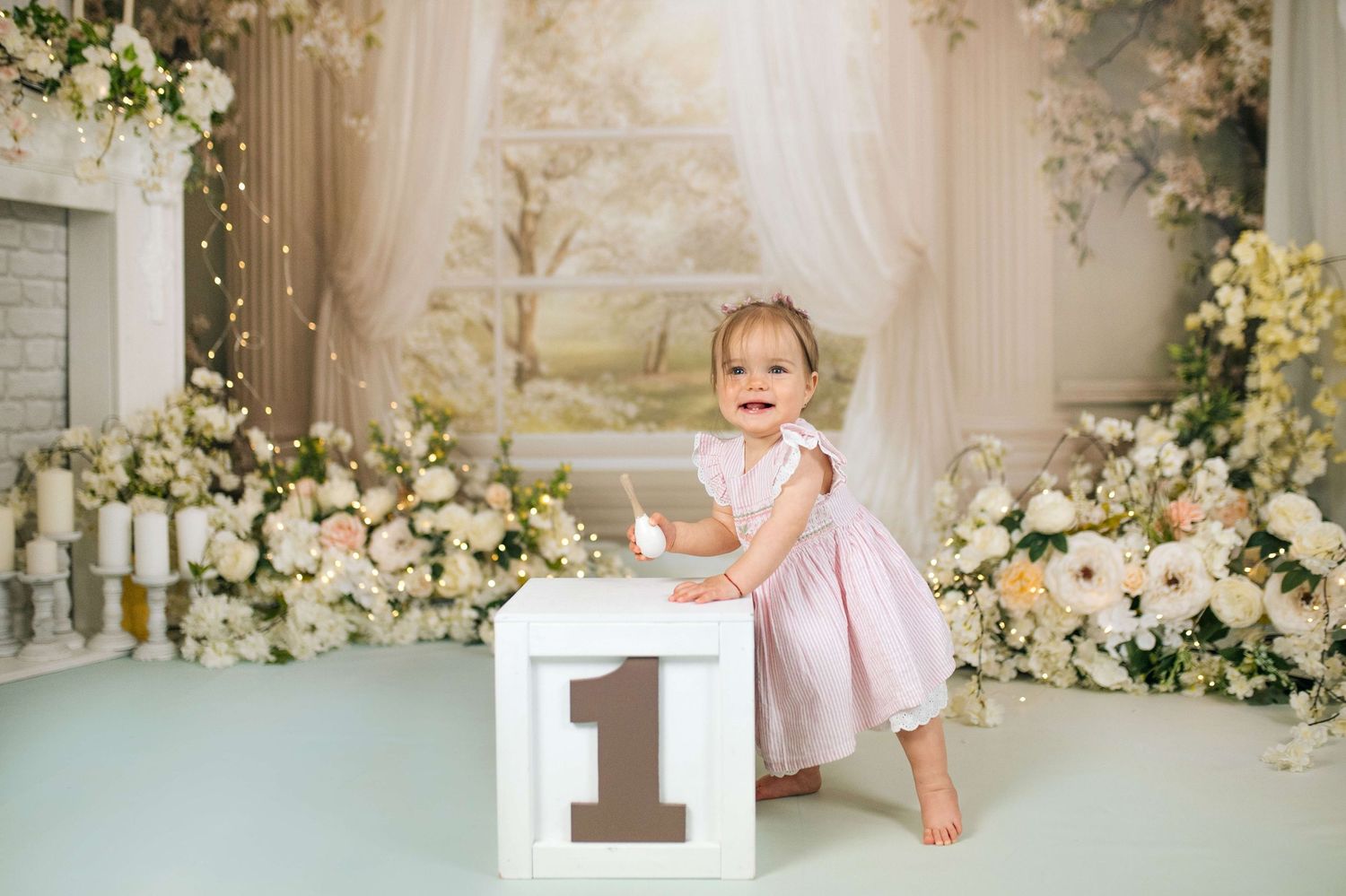 A smiling baby girl in a pink dress stands beside a number one block surrounded by flowers for her first birthday.