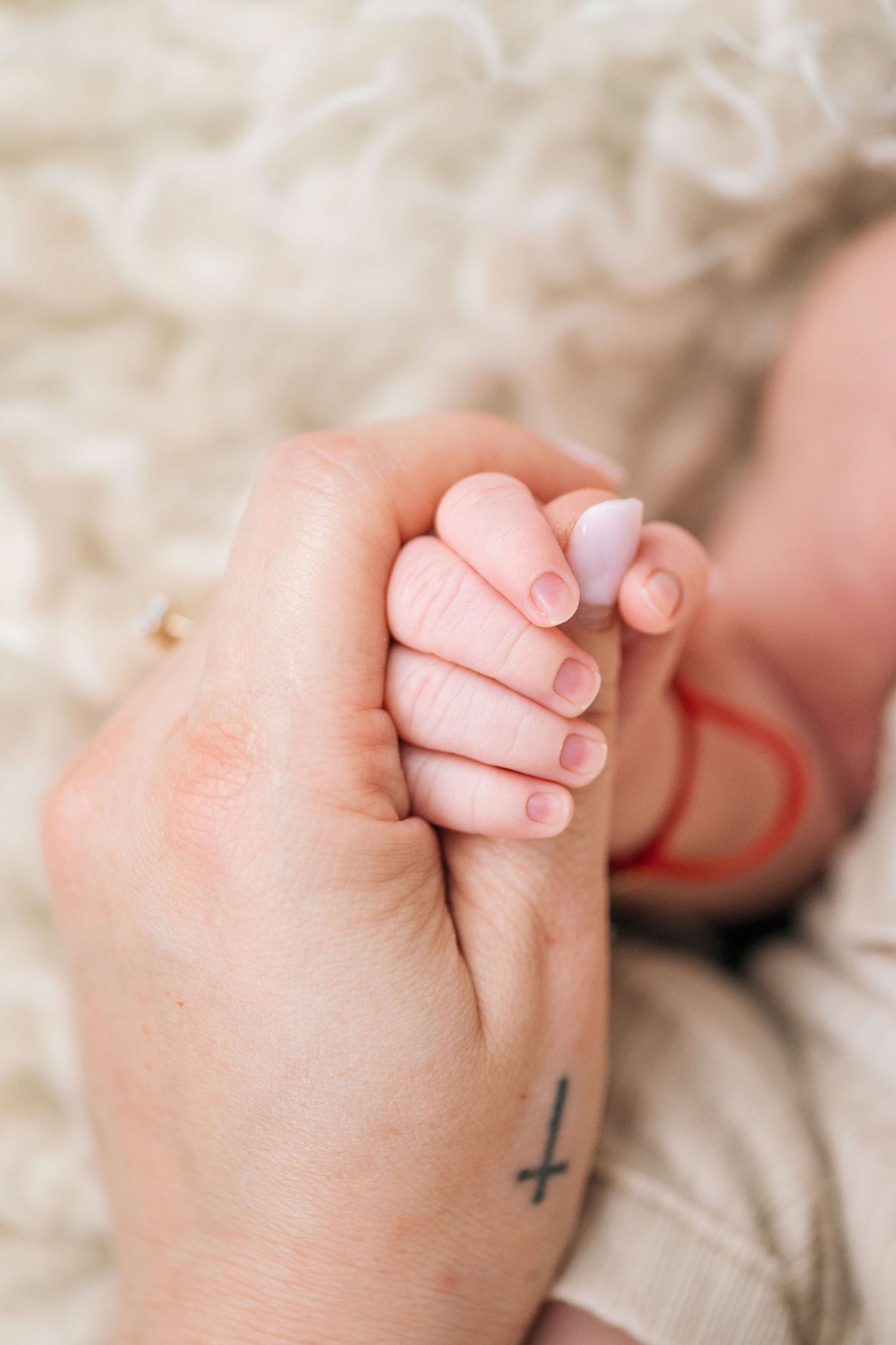 A parent's tattooed hand gently holds a sleeping newborn baby's tiny fist on a soft white blanket.