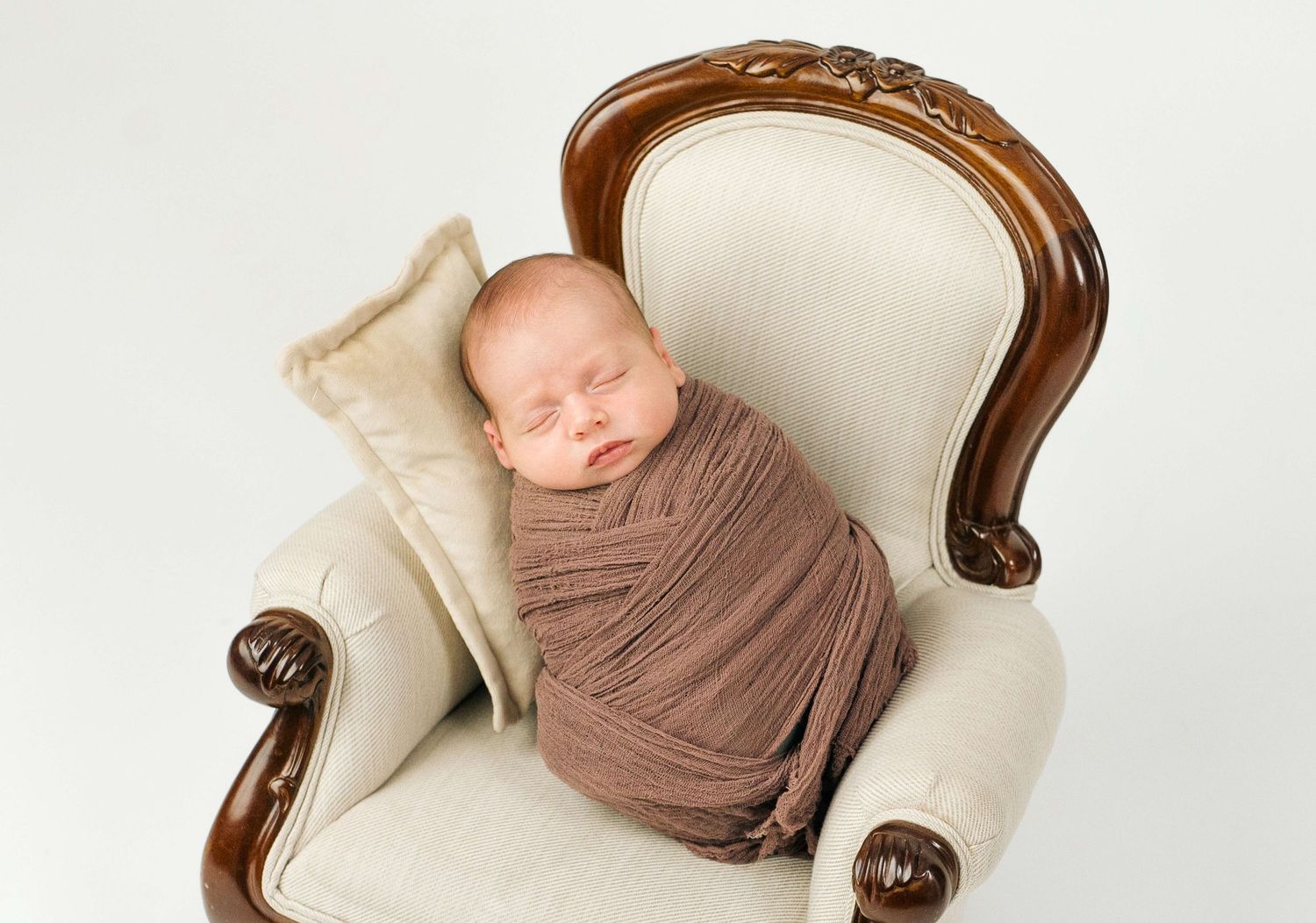 Sleeping newborn baby swaddled in brown wrap posed in a cream vintage miniature chair for a professional newborn photo.