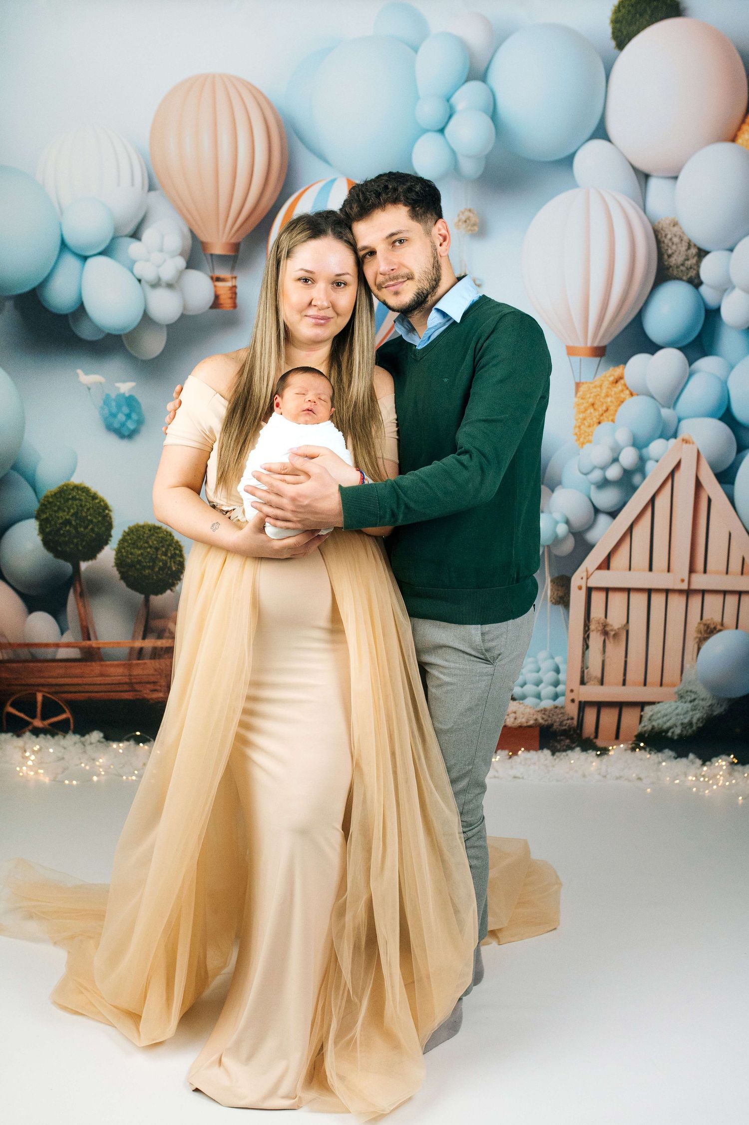 Young couple in elegant attire holds newborn baby, posing before colorful balloon and hot air balloon backdrop.