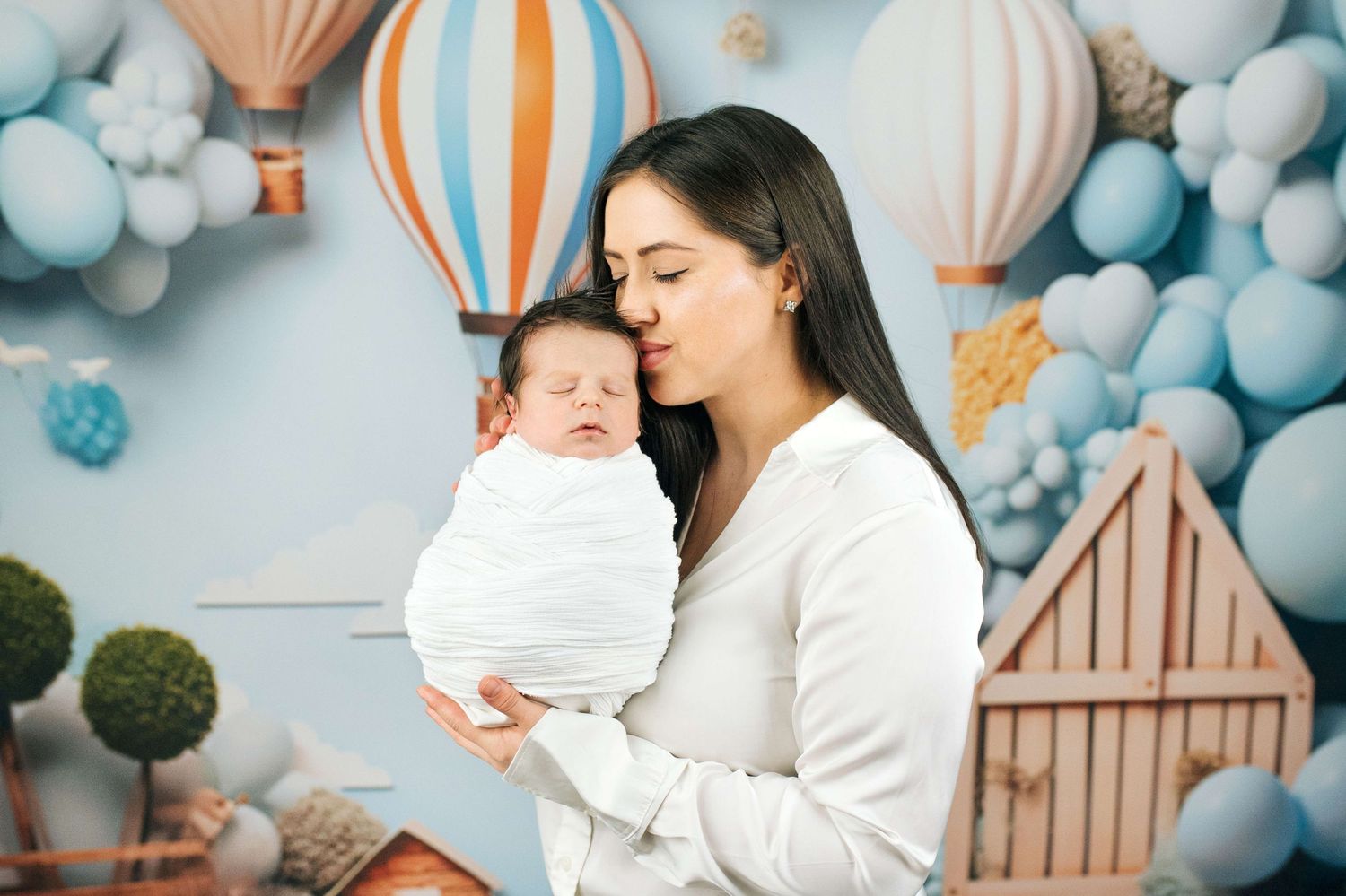 A young mother tenderly kisses her swaddled newborn baby against a pastel blue balloon backdrop.
