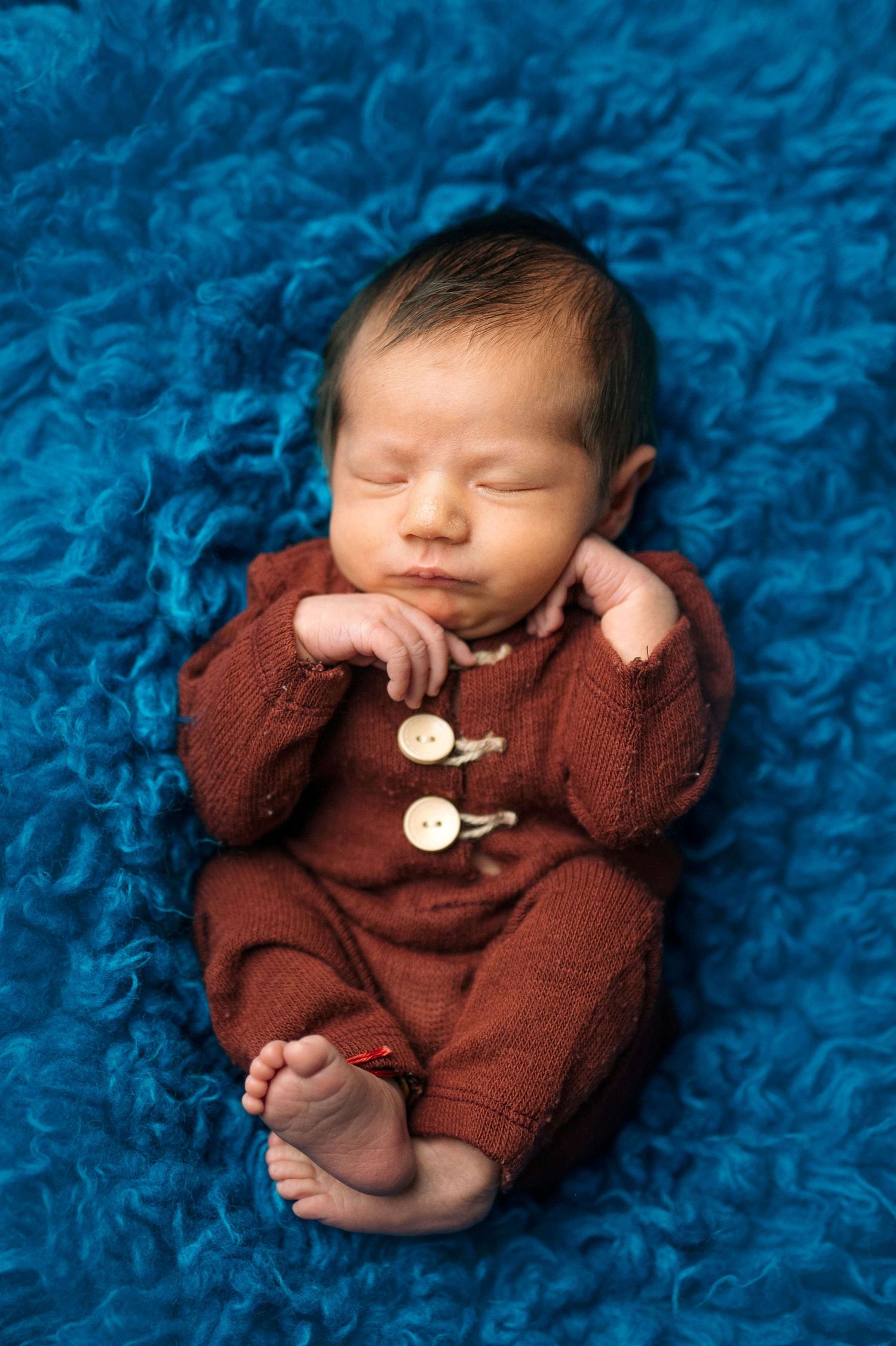 Sleeping newborn baby in red knit romper with buttons, resting on blue fluffy blanket.