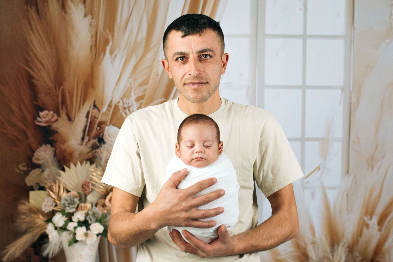 A proud father in a beige shirt gently holds his swaddled newborn baby against a floral boho backdrop.