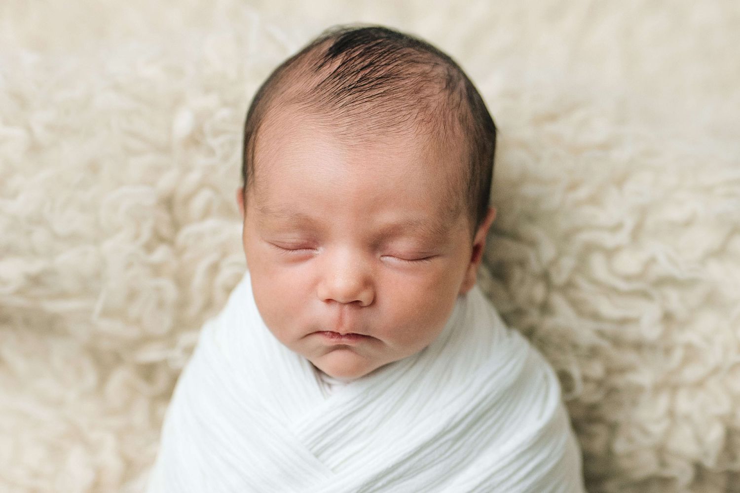A peaceful newborn baby swaddled in white wrap sleeping on a soft cream textured blanket.