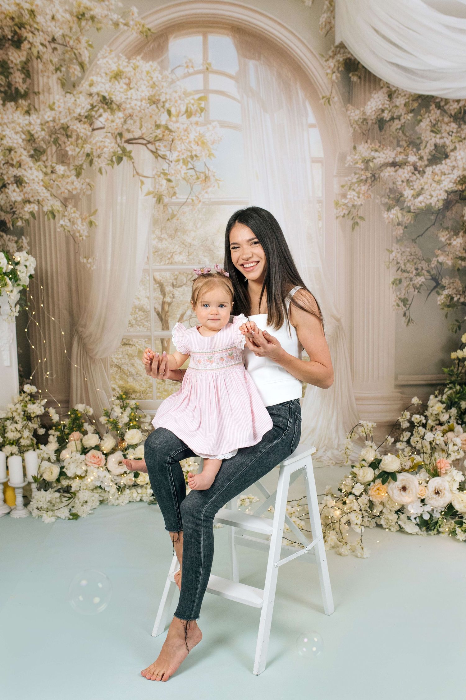A smiling mother sits on a white stool holding her toddler daughter in a pink dress, surrounded by white floral studio backdrop.