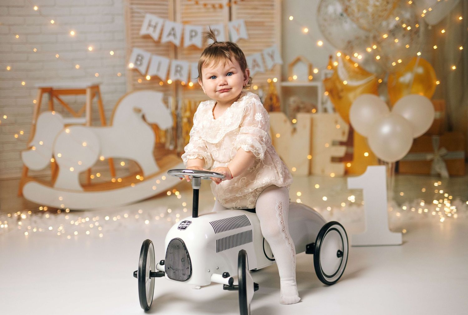 Toddler sitting on a white toy car at a first birthday party with balloons and fairy lights.