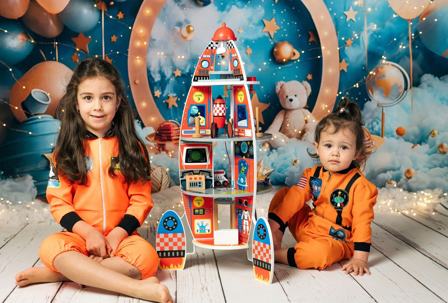 Two children in orange astronaut suits sit beside a colorful rocket toy against a space-themed backdrop.