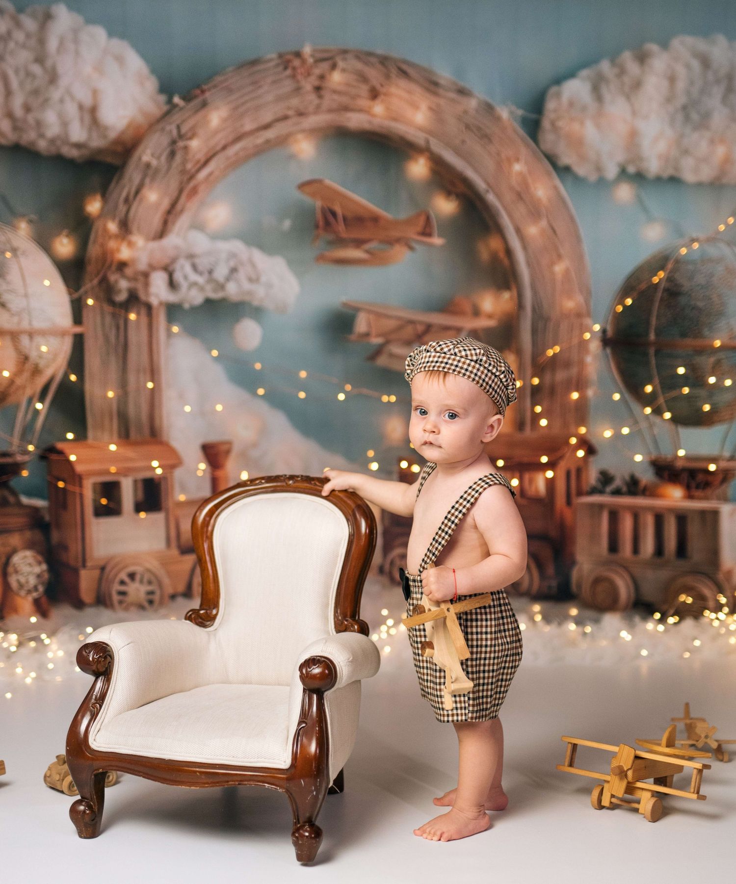 A baby boy in vintage outfit stands by a miniature chair with airplane and train themed backdrop with fairy lights.