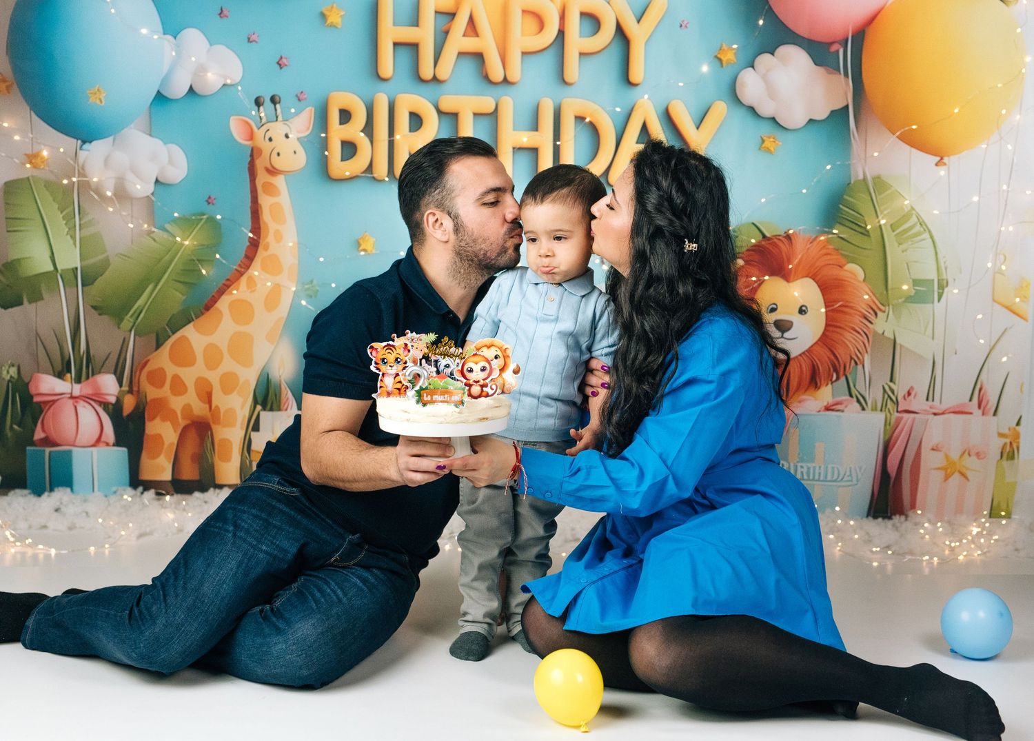Family celebrating baby's first birthday with cake, colorful jungle animal backdrop, and Happy Birthday banner.