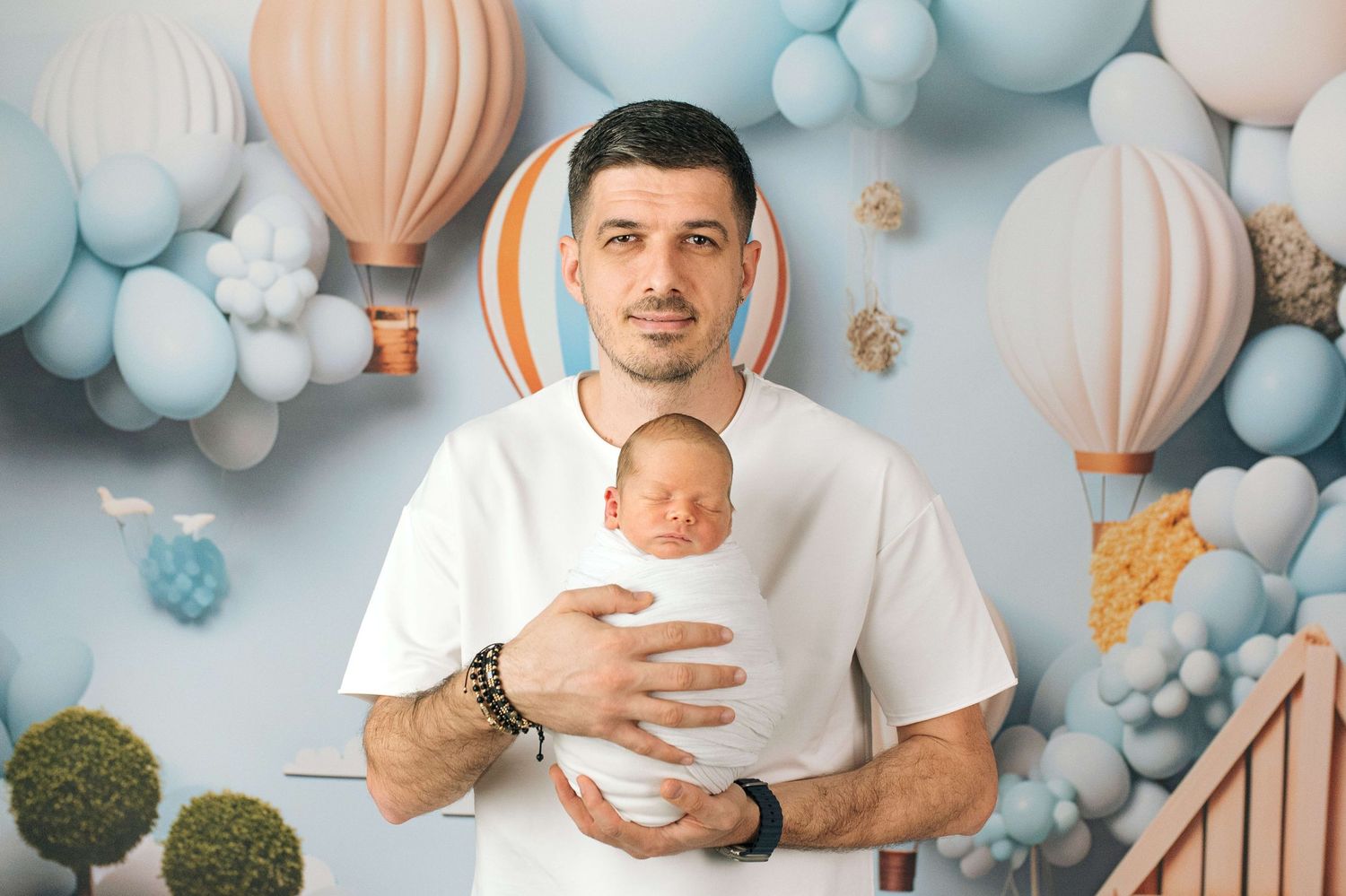 Father in white t-shirt holds sleeping newborn baby against decorative balloon backdrop.