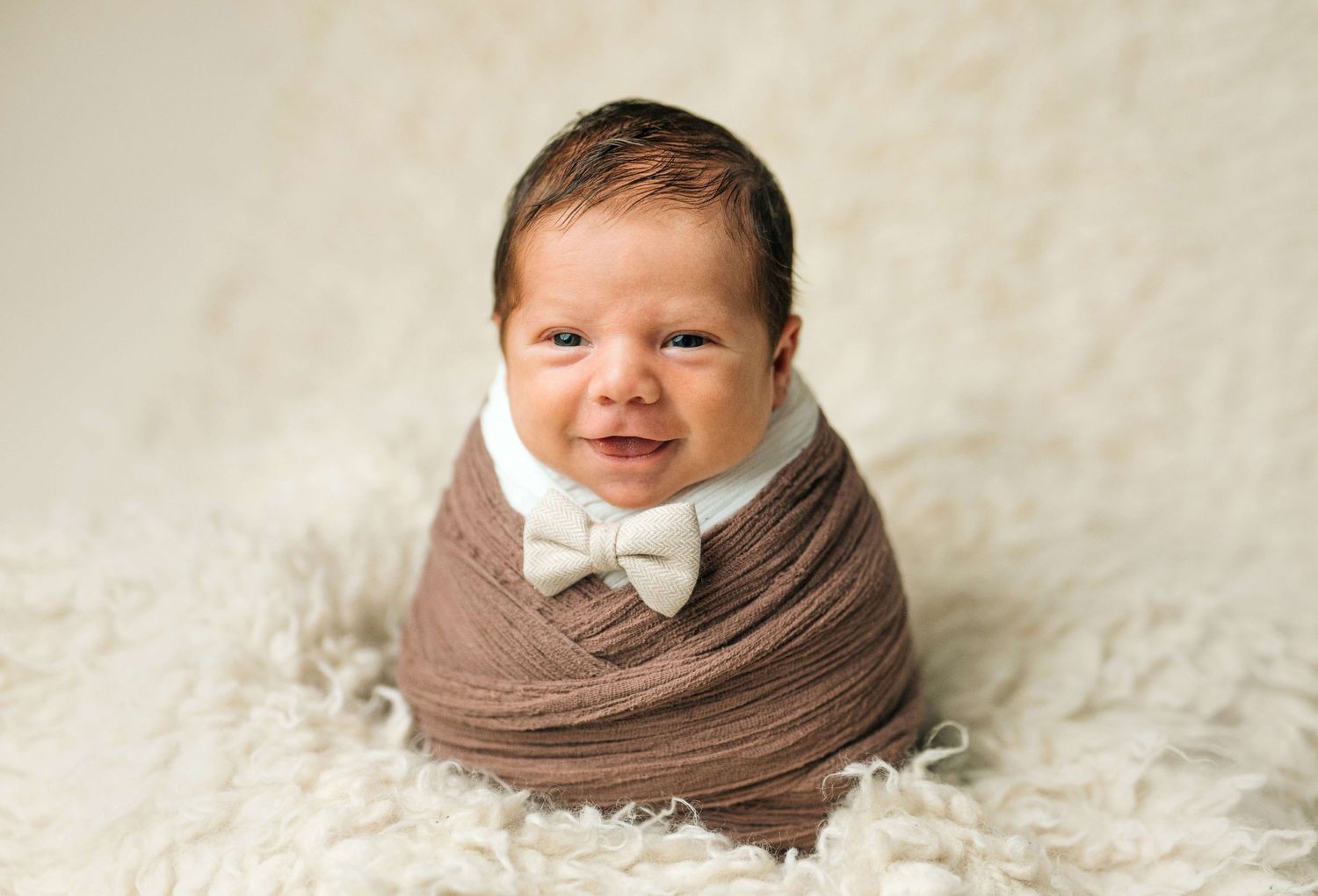 Adorable newborn baby wrapped in brown swaddle with white bow tie, smiling on fluffy white blanket.