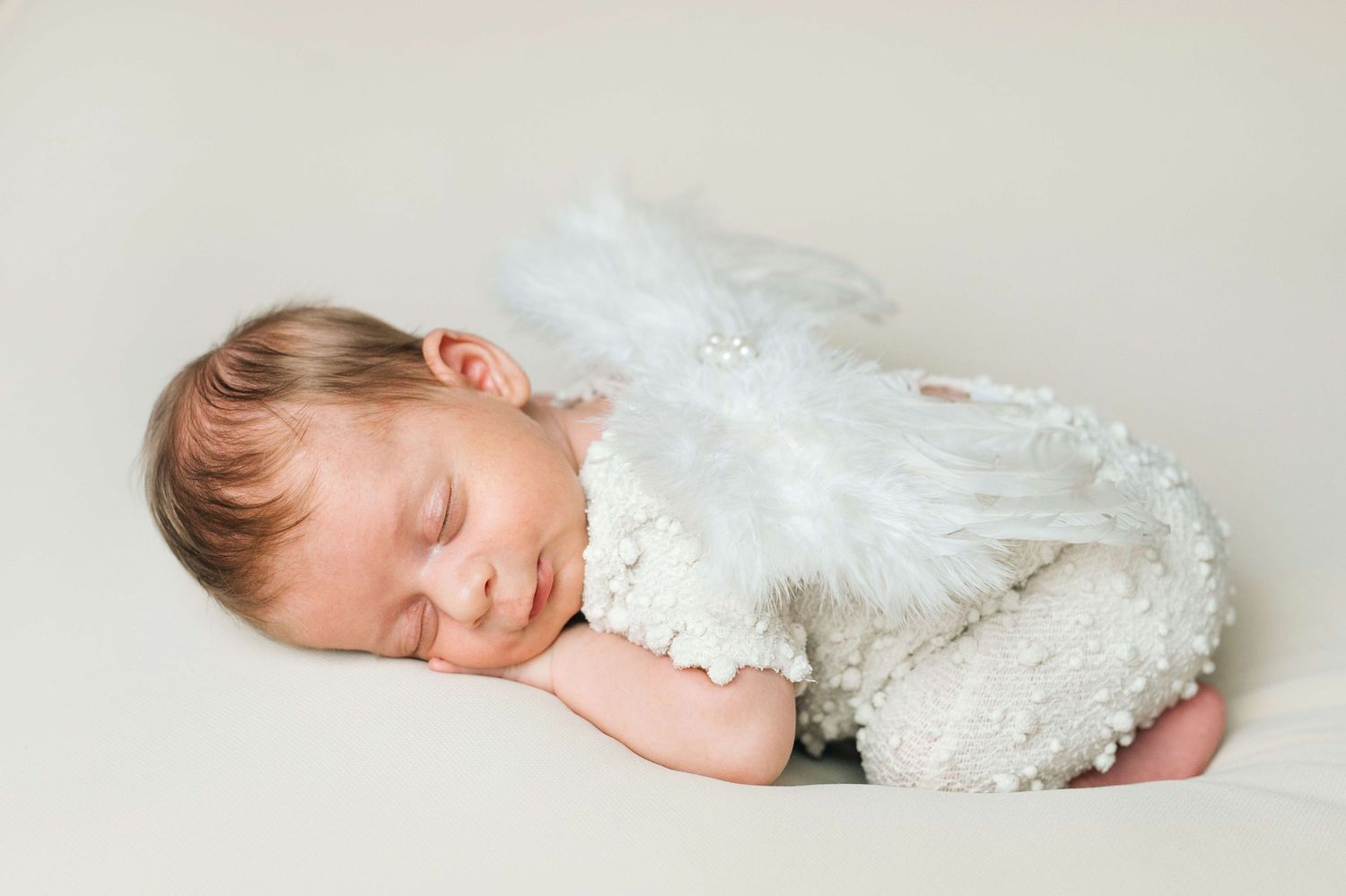 Sleeping newborn baby dressed in white feathered angel wings costume on a soft cream background.