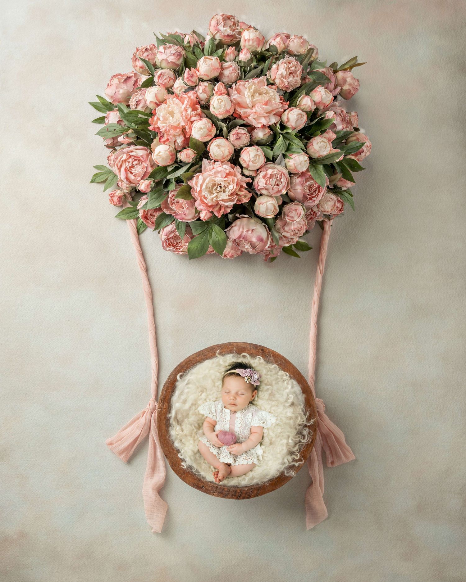 Newborn baby sleeping in a wooden bowl suspended beneath a lush pink peony hot air balloon arrangement.