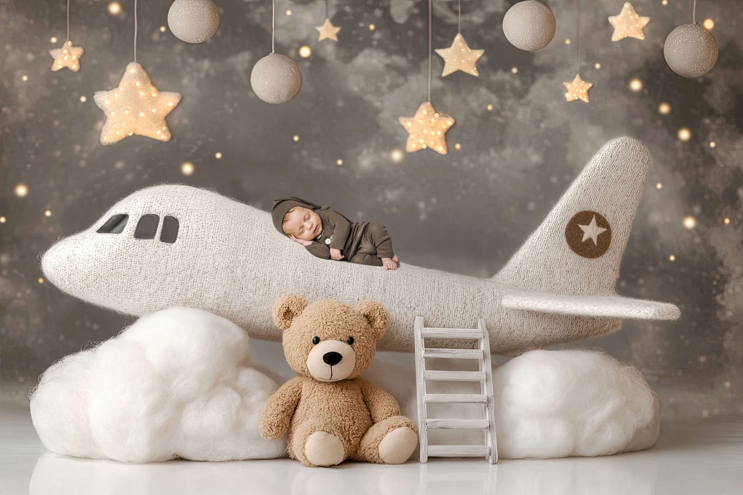 Newborn baby sleeping on a plush white airplane prop with a teddy bear, stars, and clouds in a dreamy nursery photo.