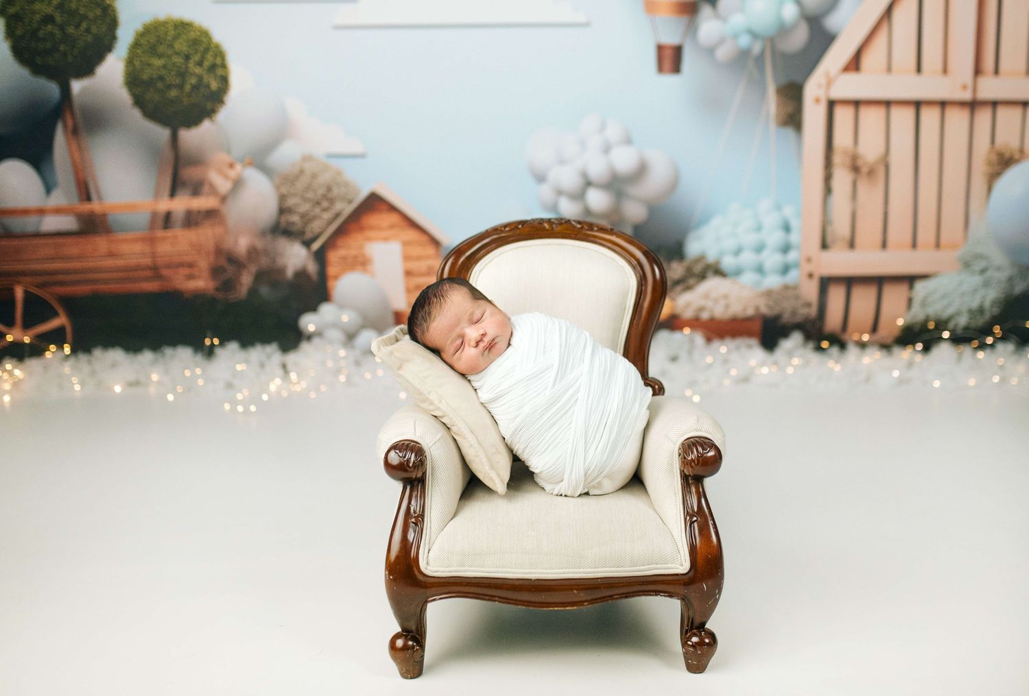 Newborn baby swaddled in white sleeping peacefully on a miniature vintage armchair in a whimsical studio setting.