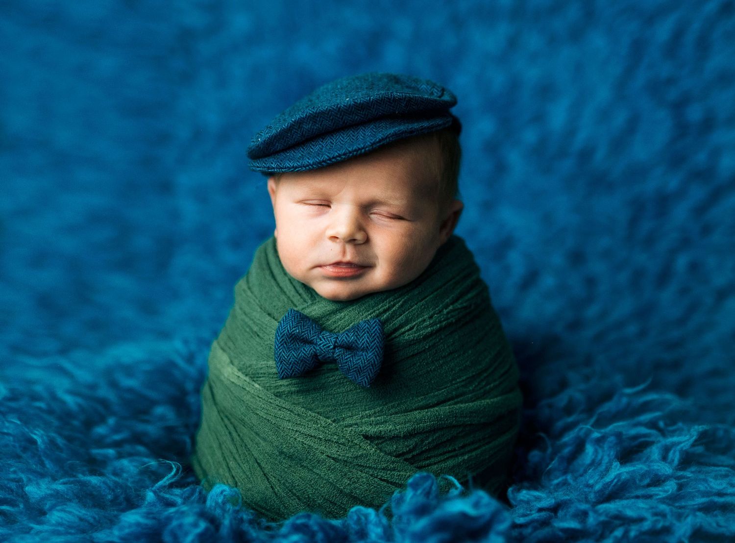 Adorable newborn baby swaddled in green wrap wearing a navy flat cap and bow tie on blue fluffy background.