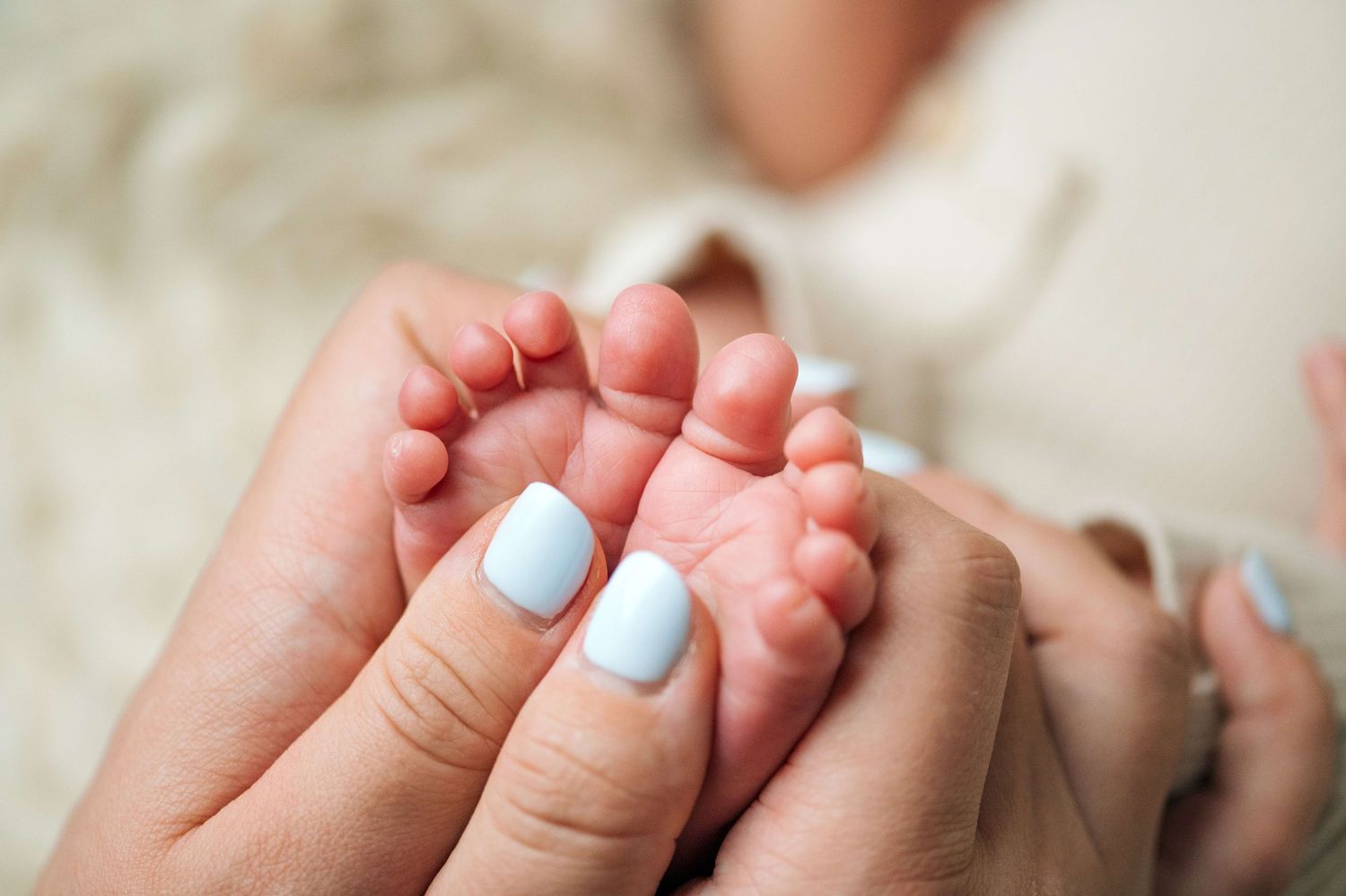 Close-up of a mother with light blue nails gently holding her newborn baby's tiny feet.