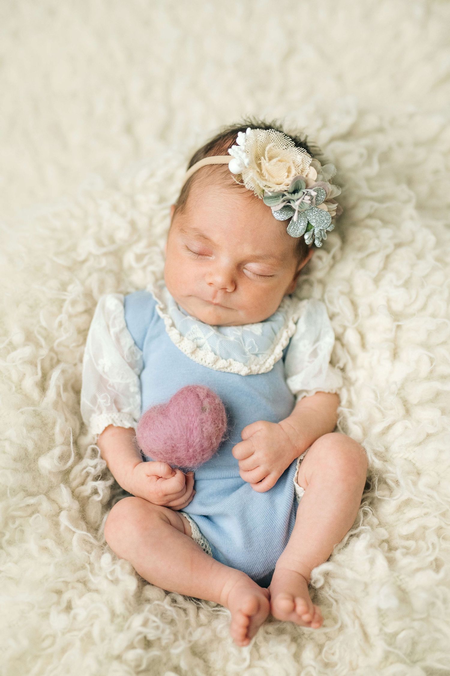 Sleeping newborn baby girl in blue romper and floral headband resting on cream fluffy blanket.