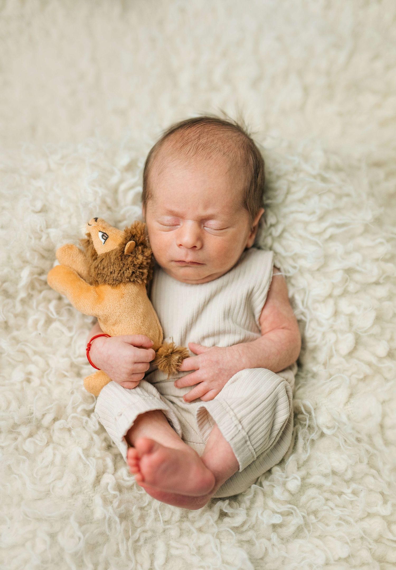 Newborn baby sleeping peacefully on white fluffy blanket, wearing cream romper, cuddling a small lion stuffed animal toy.