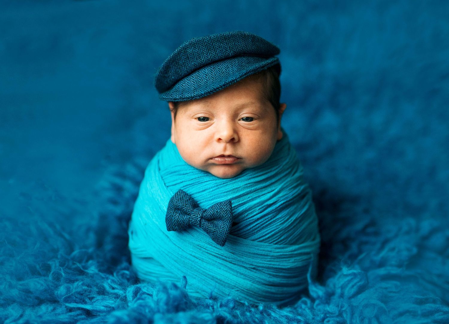 Adorable newborn baby wrapped in teal swaddle with bow tie and newsboy cap on blue fluffy blanket.