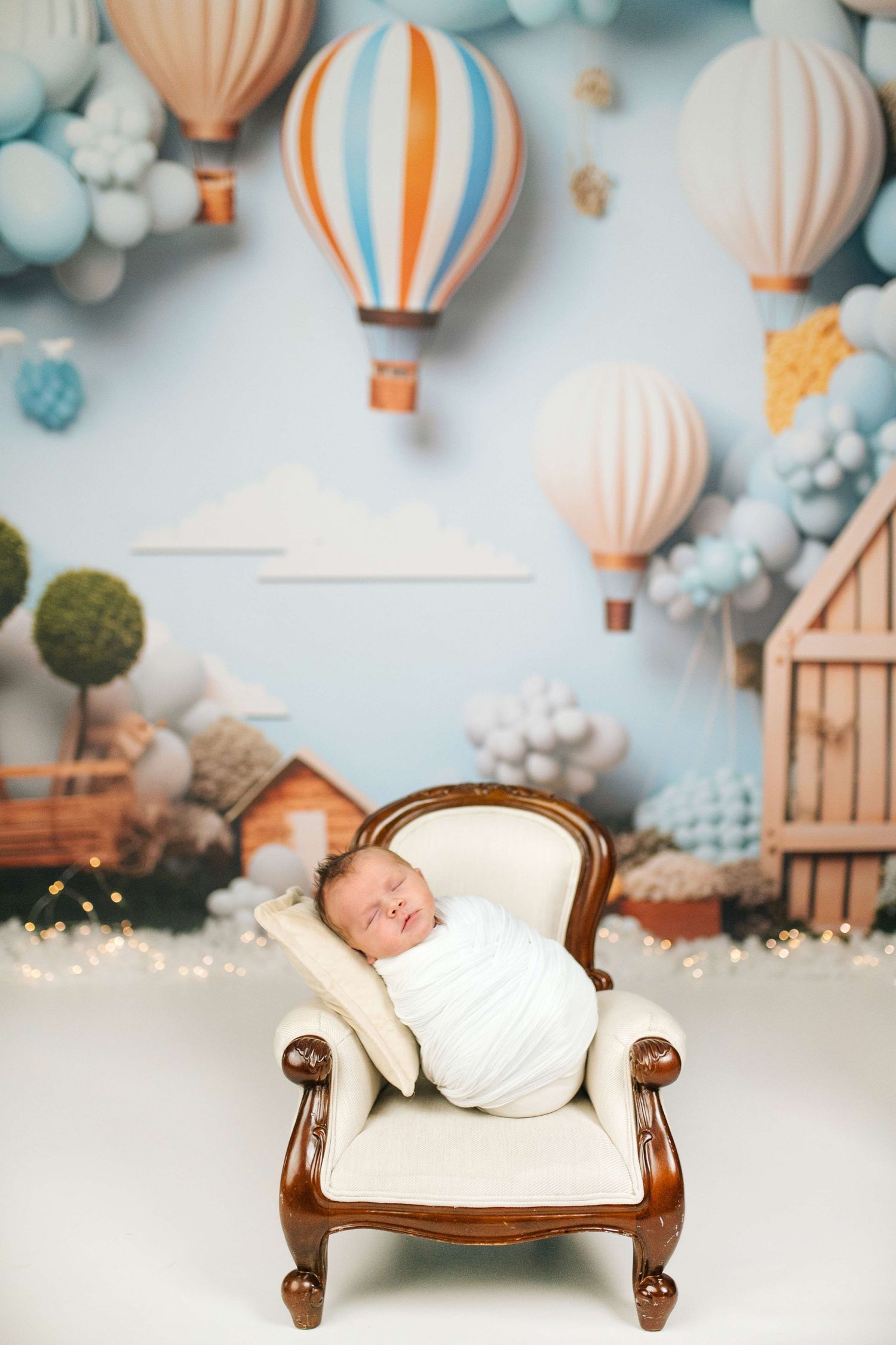 Newborn baby swaddled in white sleeping on a miniature armchair with hot air balloon nursery backdrop.