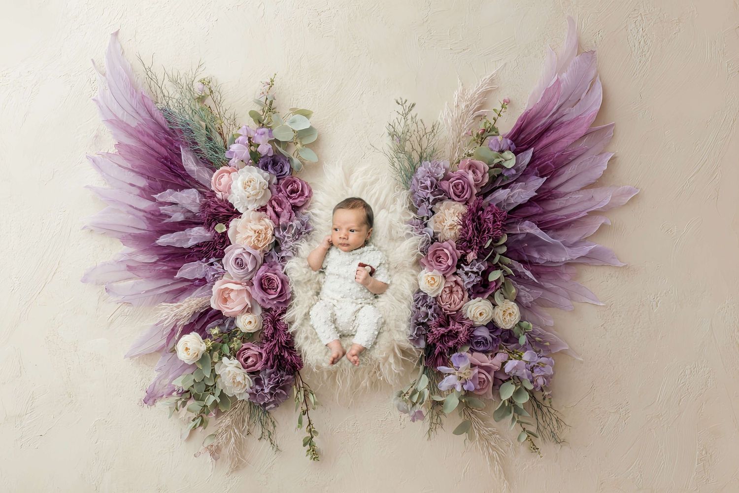 Newborn baby lying on floral angel wings backdrop with purple roses and feathers in a professional photo.