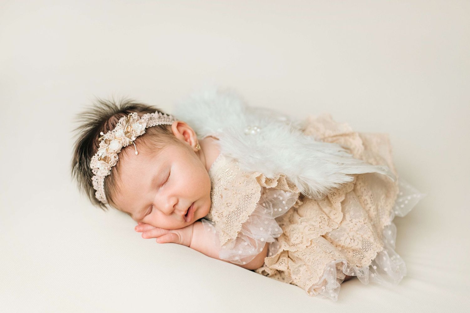 Sleeping newborn baby girl wearing a floral headband and white angel wings, wrapped in a beige lace swaddle.