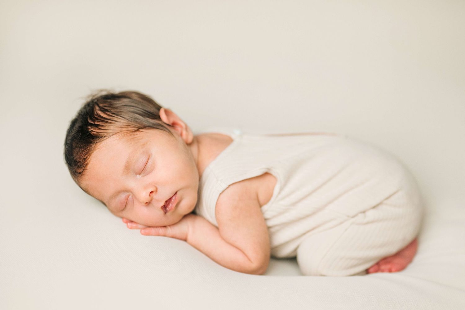 Peaceful newborn baby sleeping on white backdrop wearing cream romper, perfect newborn photography pose.