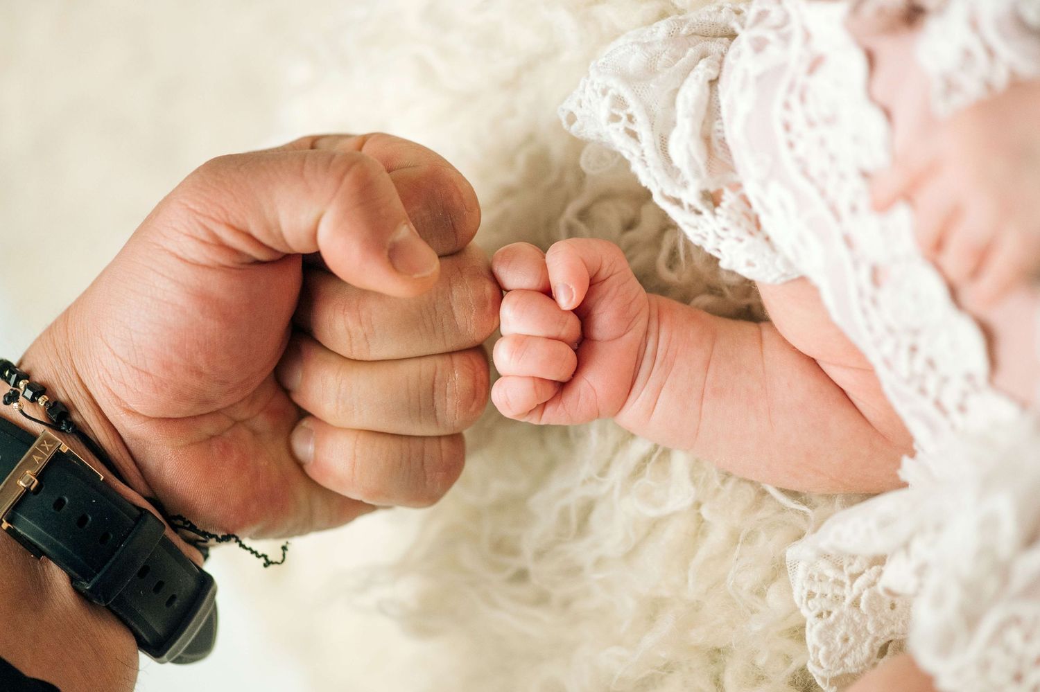 Adult fist bump with newborn baby hand, tender father-child moment in white lace setting.