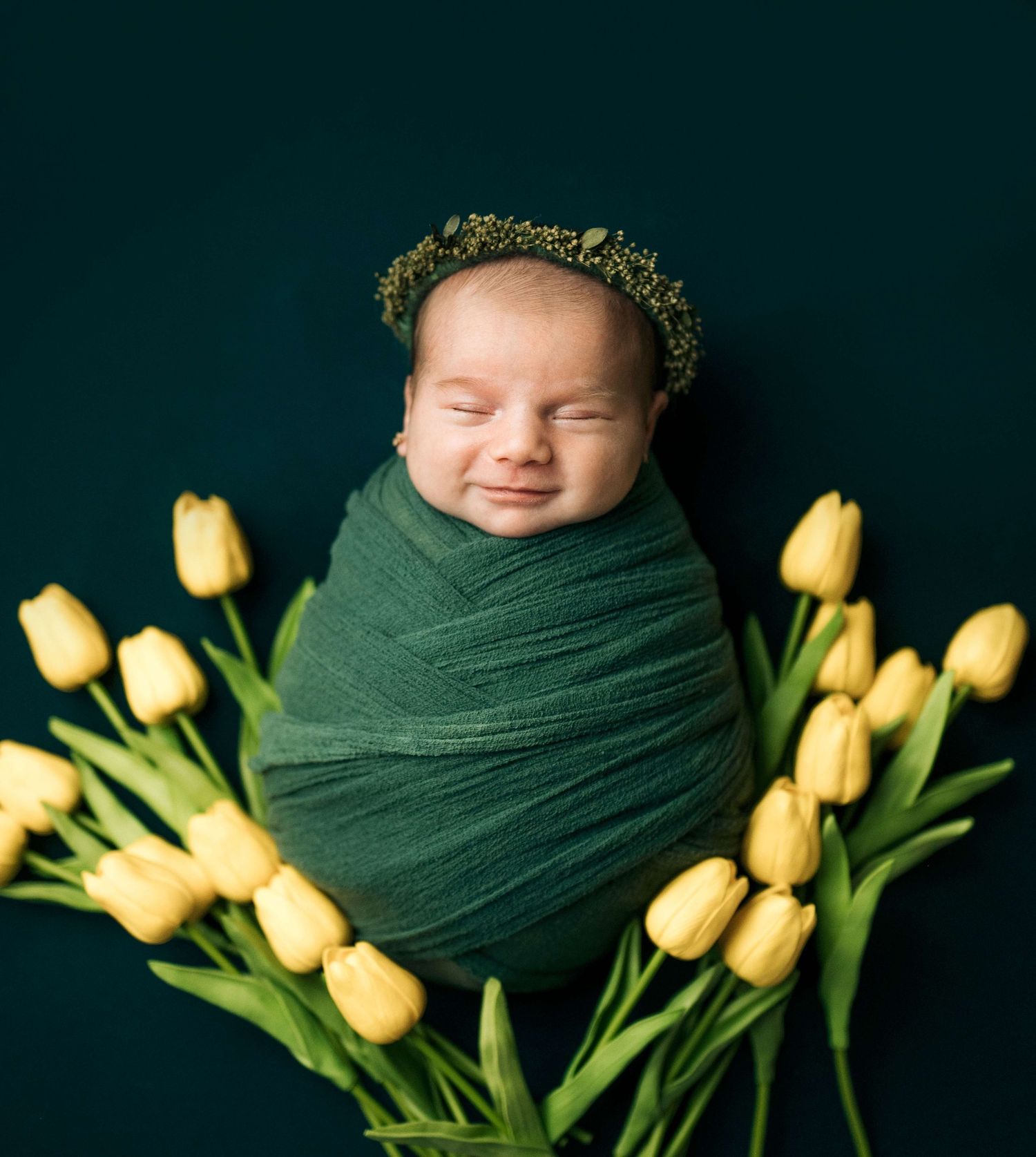 Smiling newborn baby swaddled in green wrap with floral crown, surrounded by yellow tulips on dark teal background.