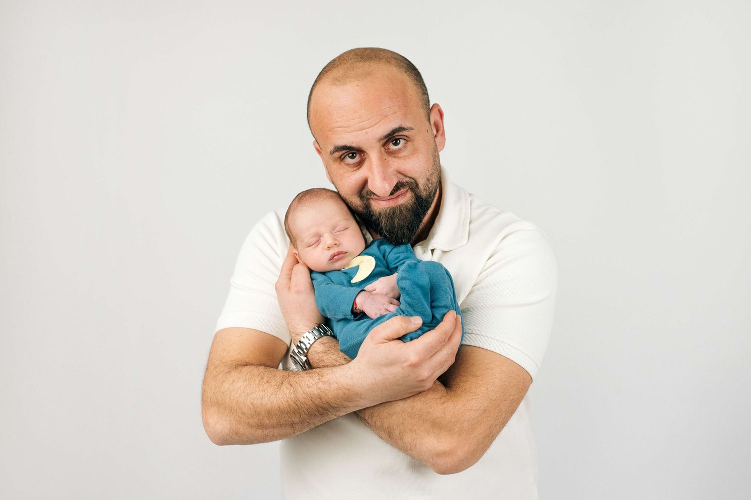 Bald father with beard proudly holds sleeping newborn baby in blue outfit against white background.