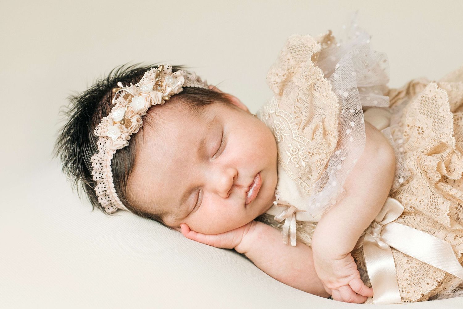 Sleeping newborn baby girl in gold lace outfit and floral headband posed on cream background.