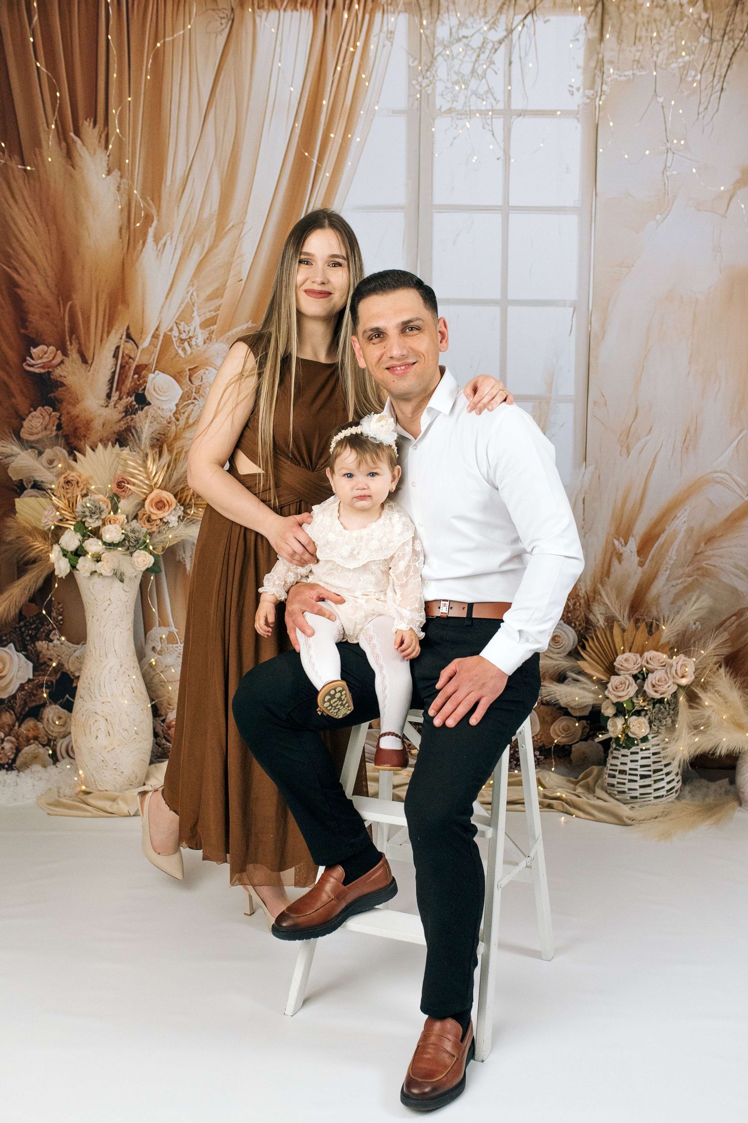A happy young family poses in a boho-styled studio, with mom in brown dress, dad in white shirt, holding their baby girl.