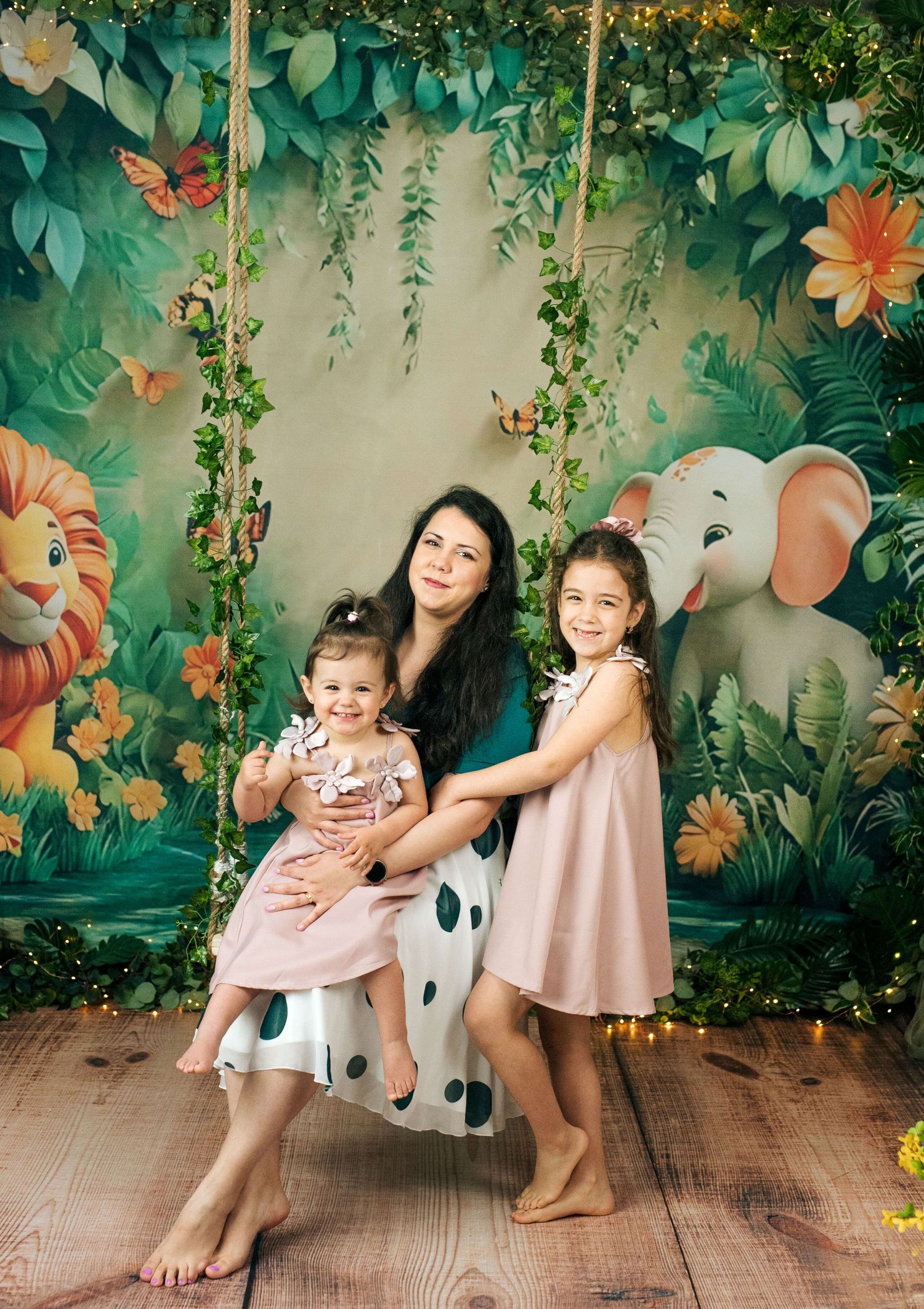 A mother sits on a jungle-themed swing with two young daughters in pink dresses, surrounded by tropical foliage and animal artwork.