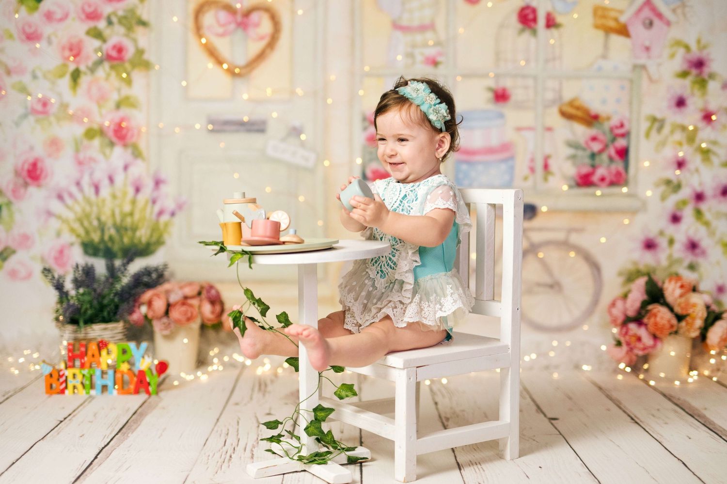 Baby girl in gray romper sits at white table with birthday cake, surrounded by floral decorations and fairy lights.
