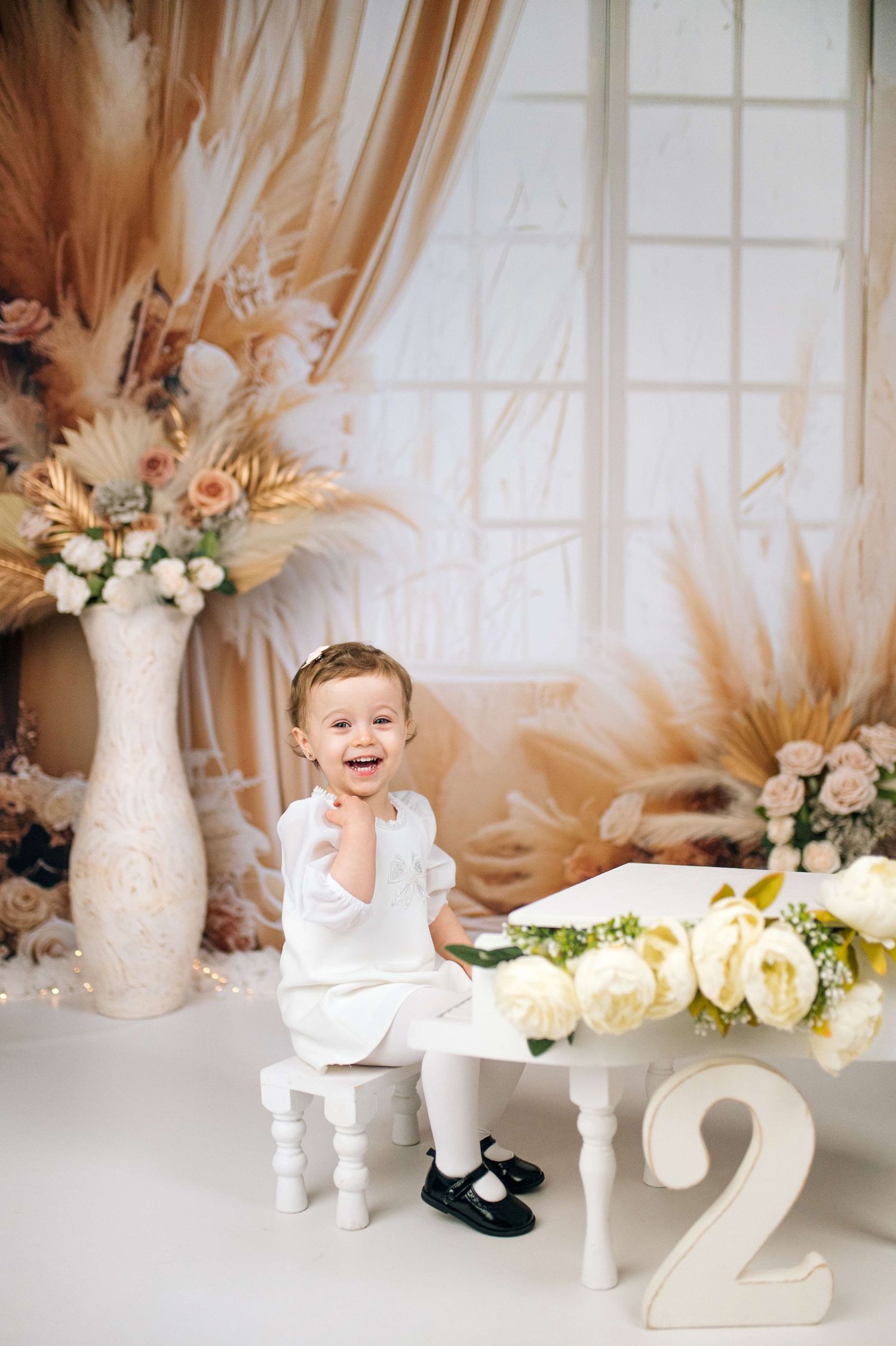 Smiling toddler in white outfit sits by a table with roses and a number 2, surrounded by floral decor.