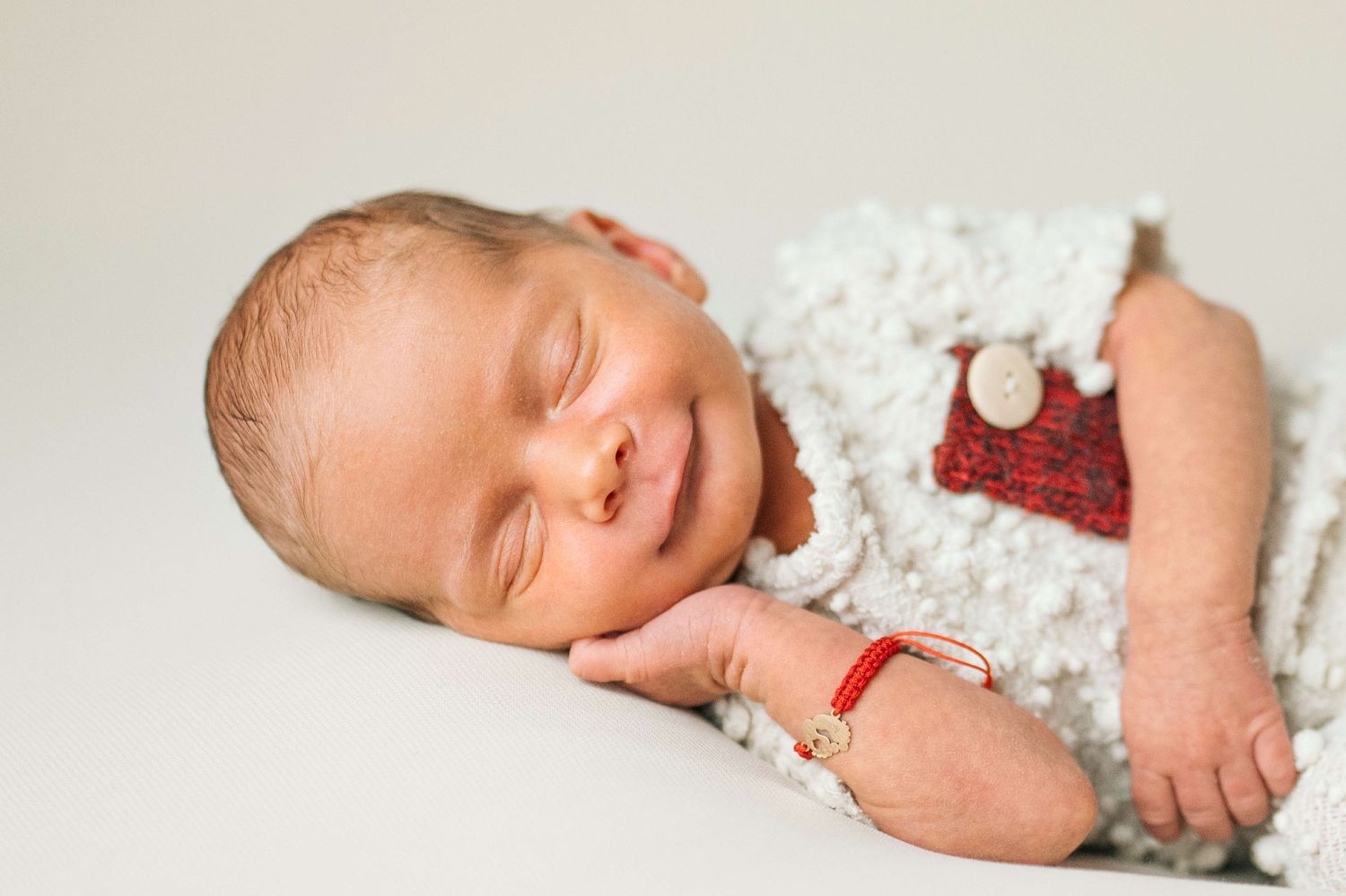 Smiling newborn baby sleeping peacefully in white fluffy outfit with red holiday accessories and bracelet.