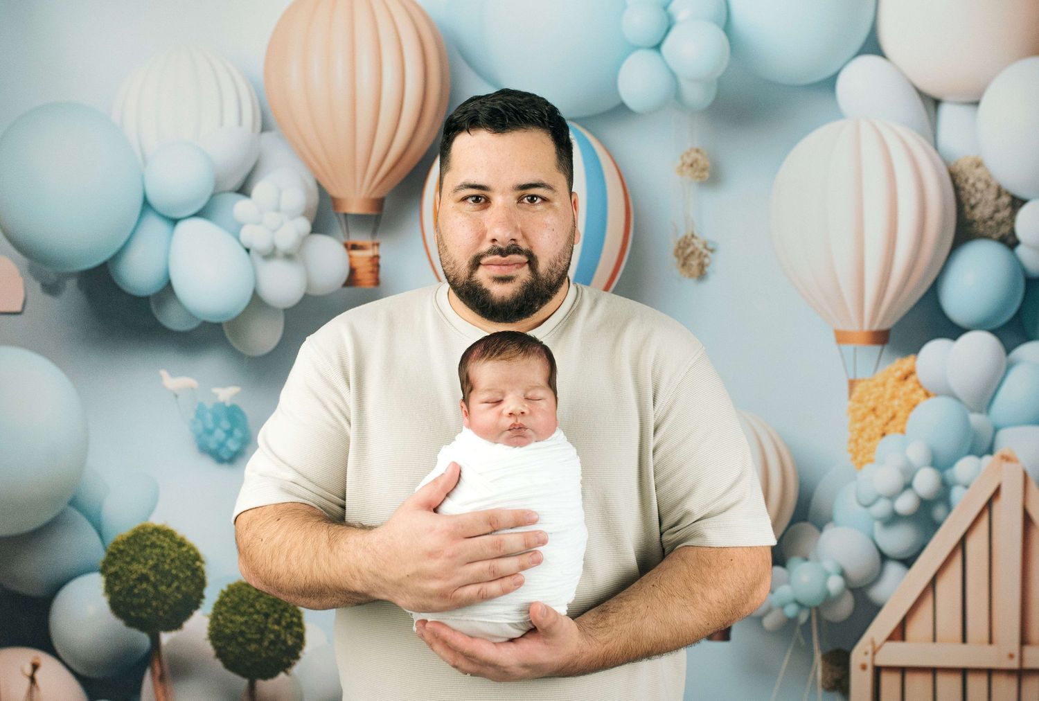 Man holding sleeping newborn baby in white swaddle against blue balloon and hot air balloon backdrop.