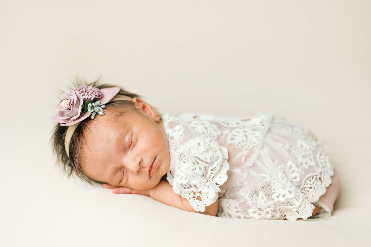 Sleeping newborn baby girl wrapped in white lace, wearing a floral headband, posed on a cream background.