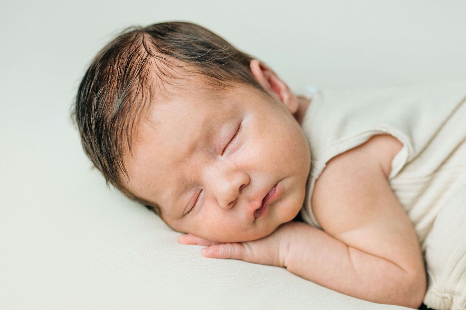 Peaceful newborn baby sleeping on a soft white blanket, with dark hair and tiny hands tucked under cheek.