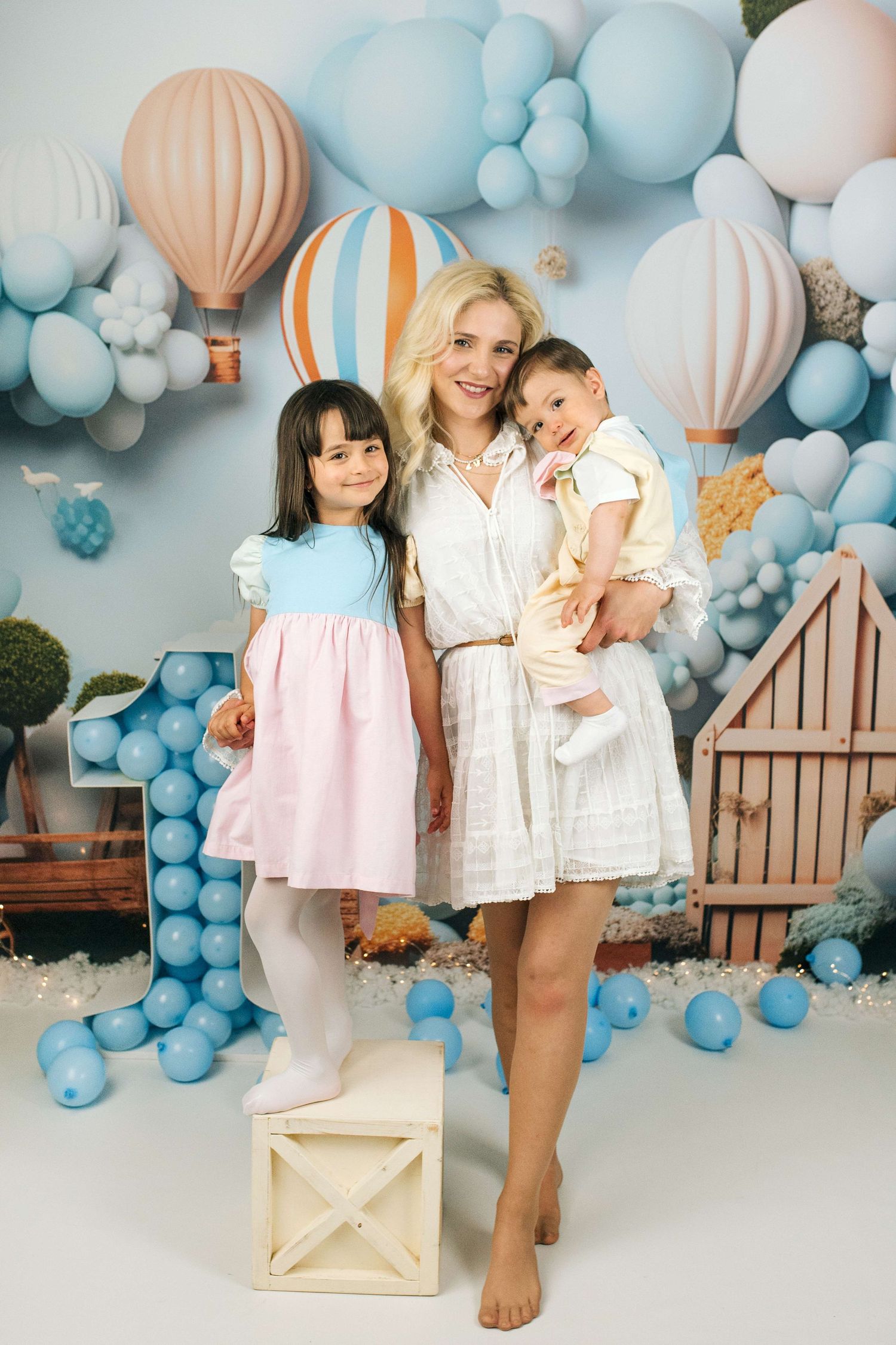 A smiling blonde woman holds a toddler while posing with a dark-haired girl in a balloon-themed birthday photo setup.