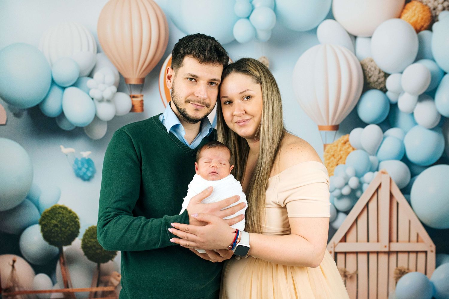 A couple poses with their newborn baby in front of a blue balloon and hot air balloon backdrop.