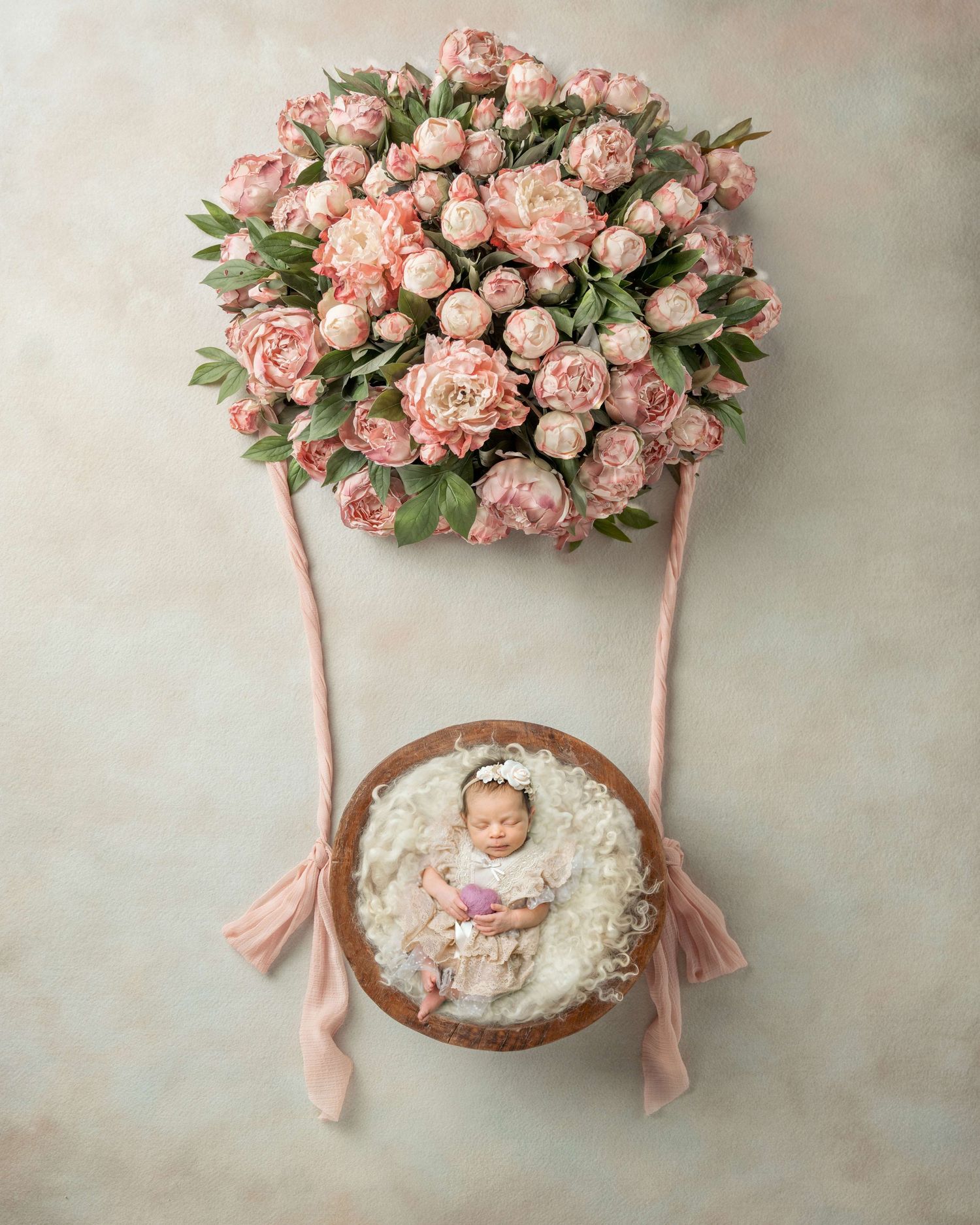 Newborn baby sleeping in a wooden bowl suspended from a pink floral hot air balloon with peony flowers.