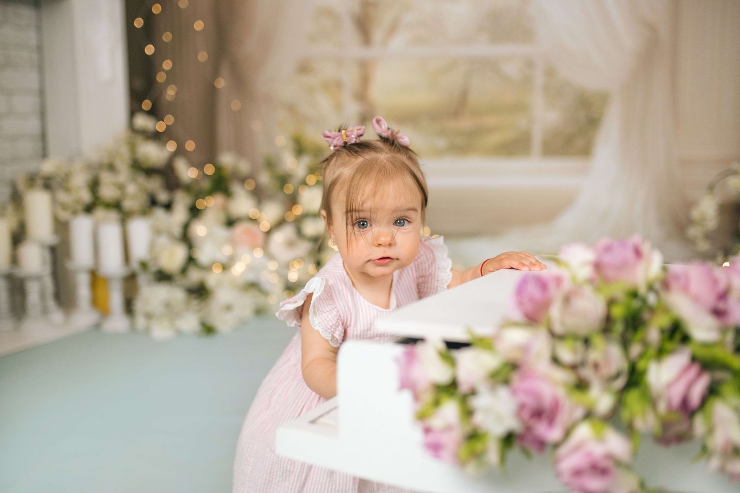 Adorable baby girl in pink dress with floral headband standing by a white piano surrounded by flowers.