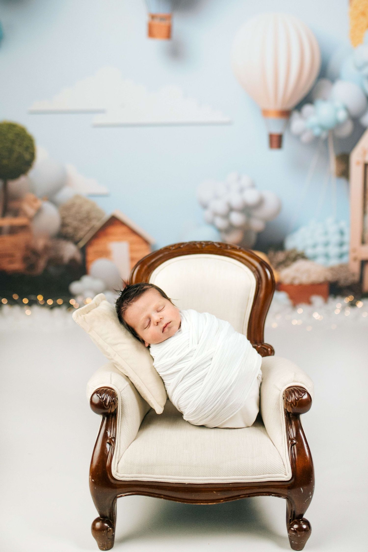 Newborn baby swaddled in white wrap sleeping peacefully on a miniature cream armchair with hot air balloon backdrop.