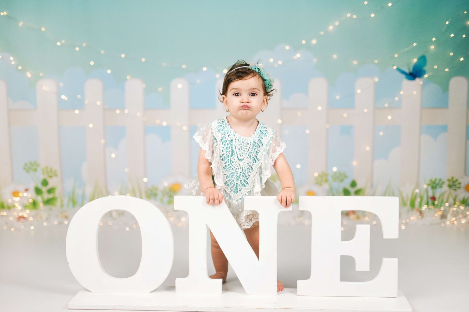 Baby girl stands behind white ONE letters for her first birthday photo session with fairy lights and pastel backdrop.