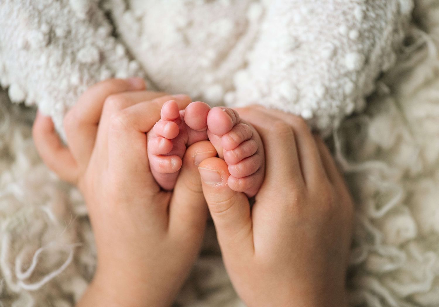 Adult hands gently cradling tiny newborn baby feet on a soft white blanket.
