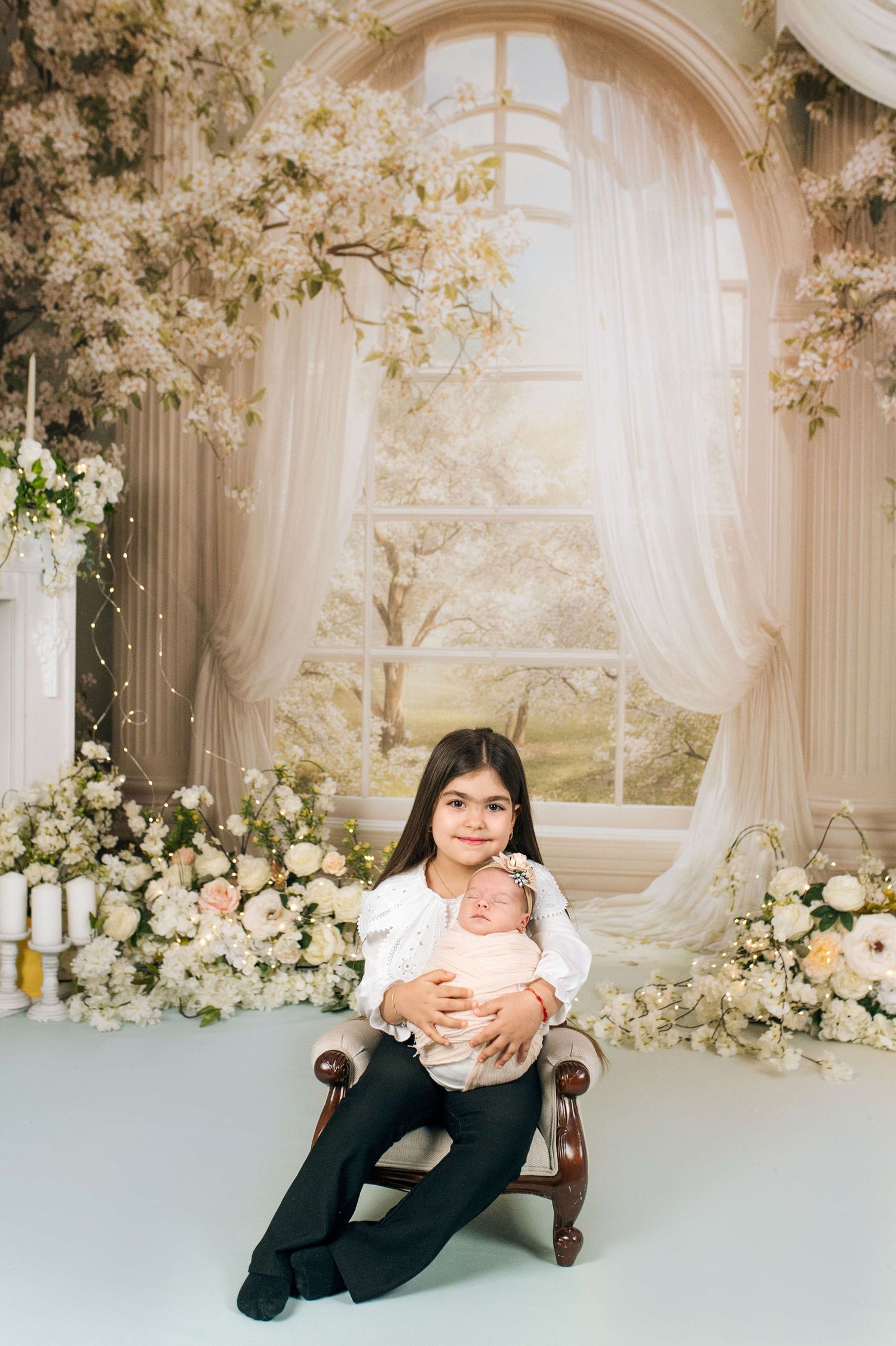 Young girl sits on chair holding sleeping newborn baby, surrounded by white floral backdrop and elegant curtains.