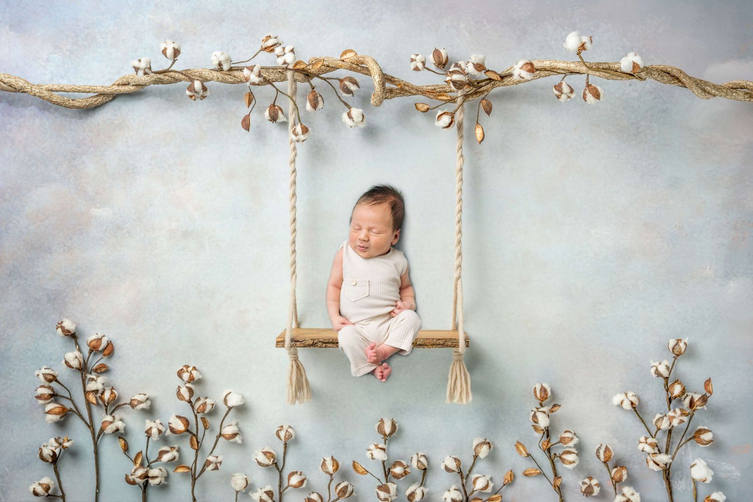Sleeping newborn baby in white outfit on wooden swing surrounded by cotton branches on soft blue background.